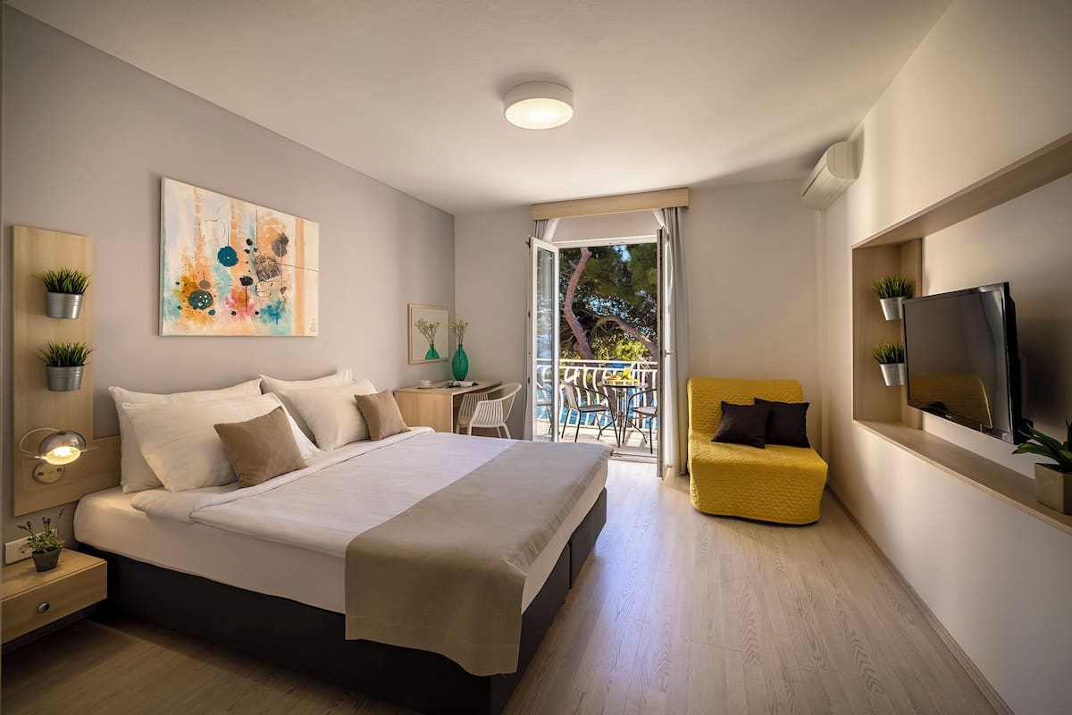 A modern bedroom with a large bed, a yellow armchair, a TV, and a balcony with a view.