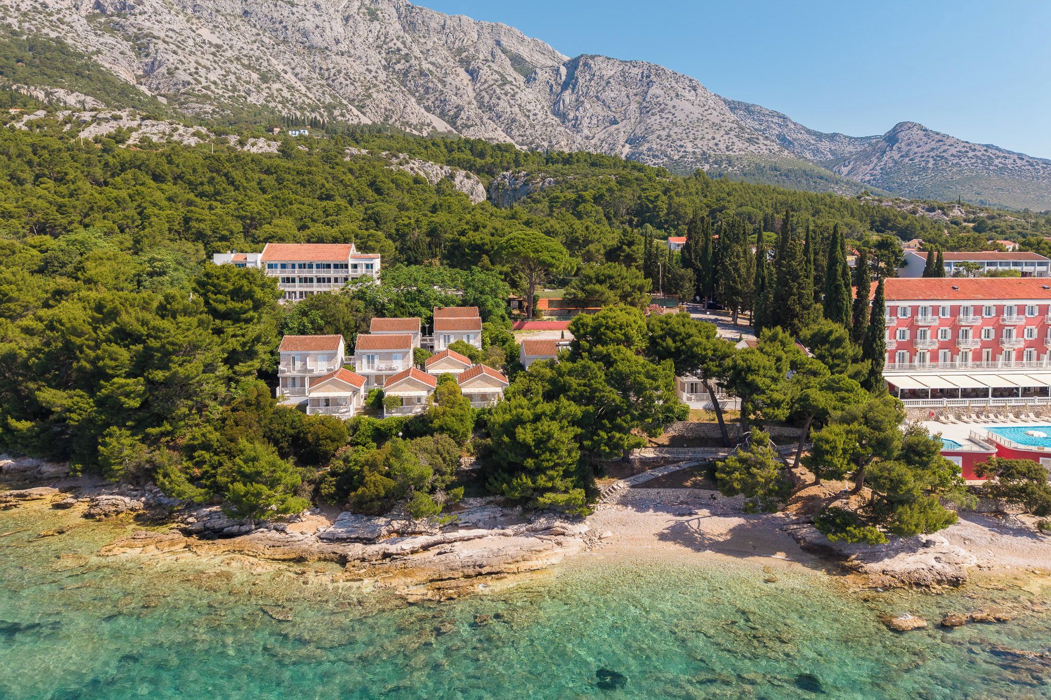 A coastal resort with multiple buildings, clear turquoise waters, and a mountainous backdrop.