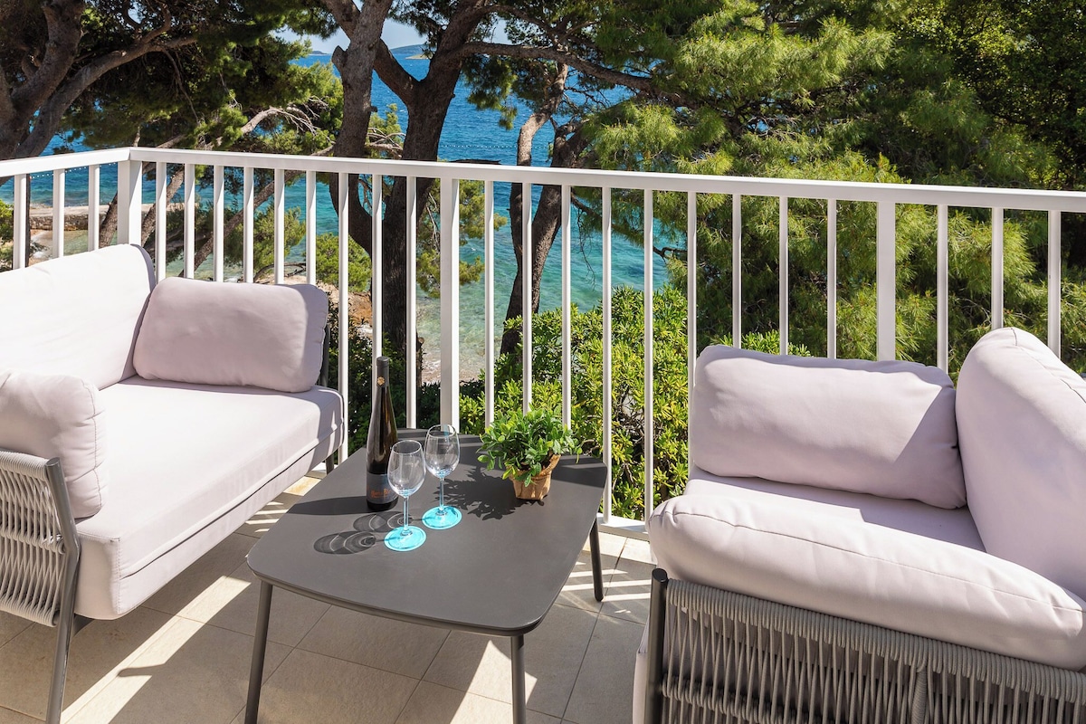 A balcony with a white railing, two white sofas, a small table with a bottle and glasses, and a view of the sea and trees.