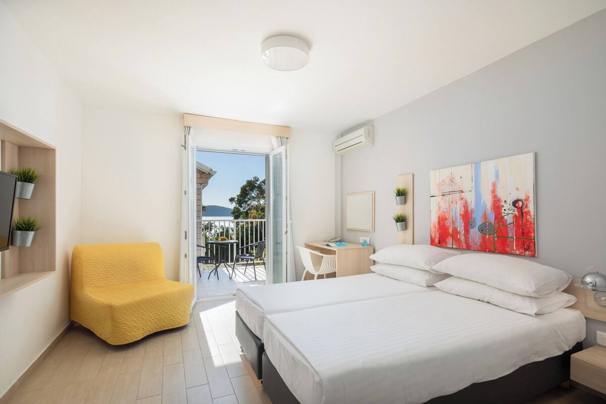 A modern hotel room with a large bed, a yellow armchair, a desk, and a balcony with a view.