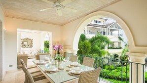 Un comedor con una mesa de vidrio, sillas de mimbre y vistas a un jardín exuberante y un edificio blanco con balcones.