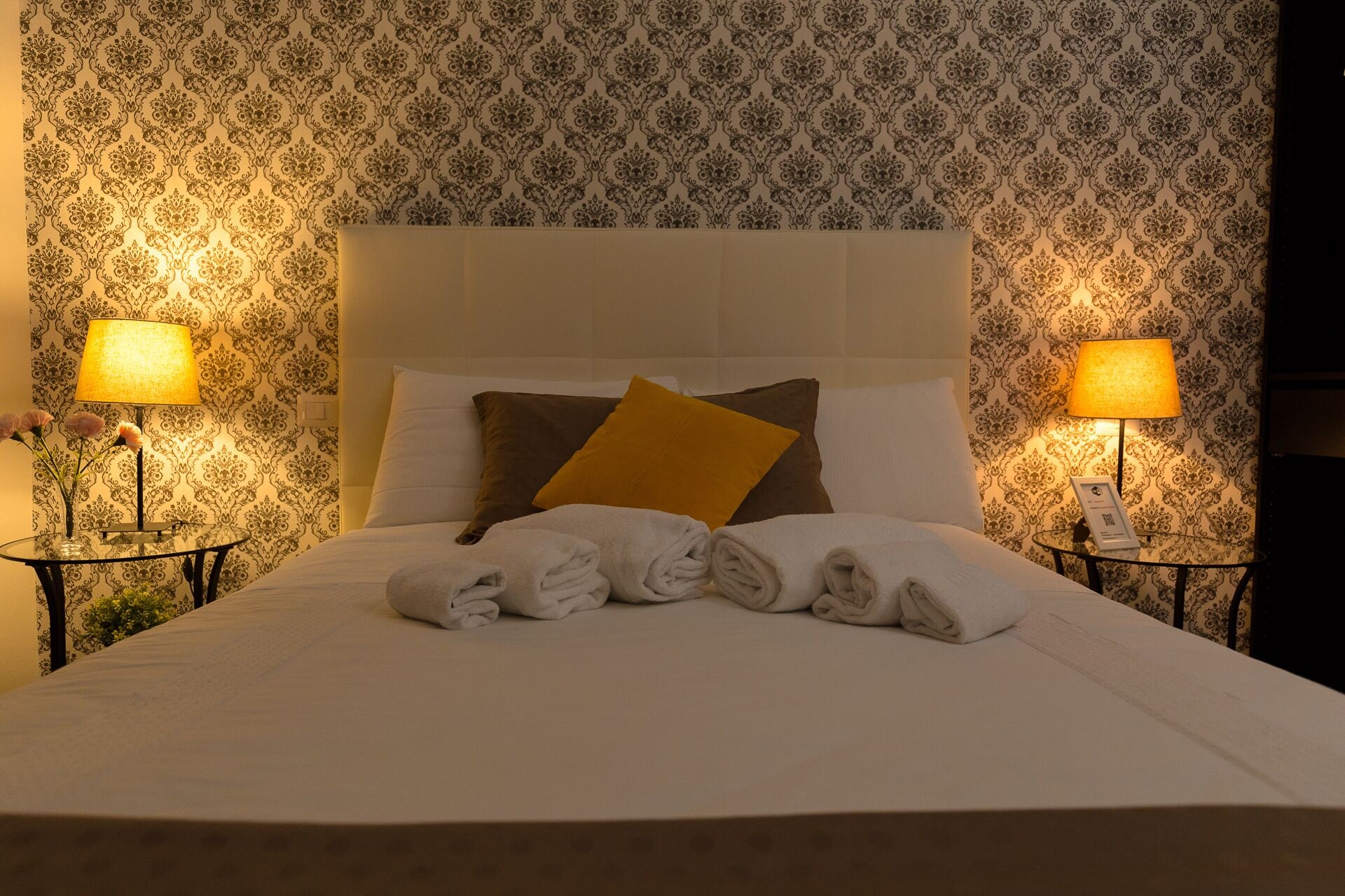 Een netjes opgemaakt bed met een wit hoofdbord, twee bedlampjes en behang met een bloemenmotief.