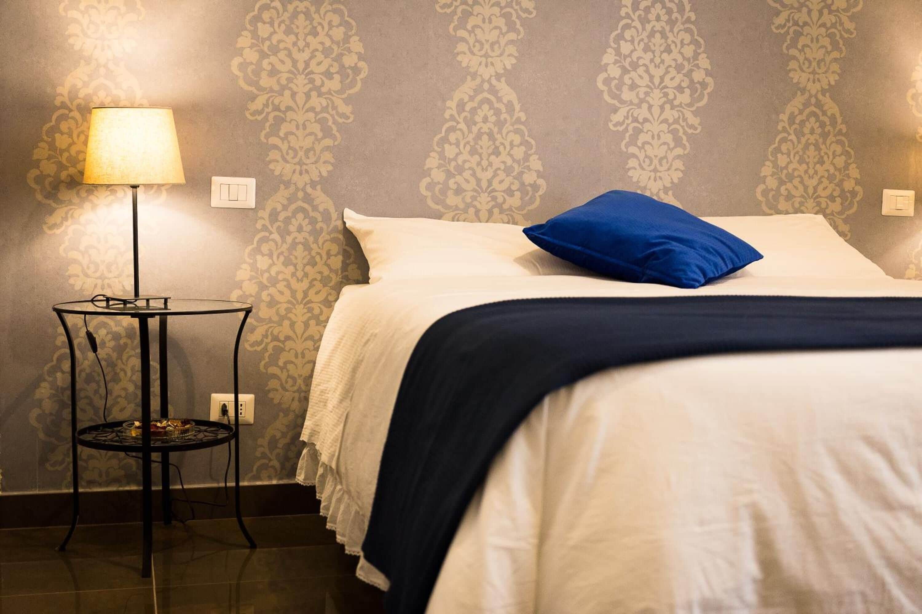 Ein ordentlich bezogenes Bett mit weißer und blauer Bettwäsche, ein kleiner runder Tisch mit einer Lampe und Wandpapier mit Muster.