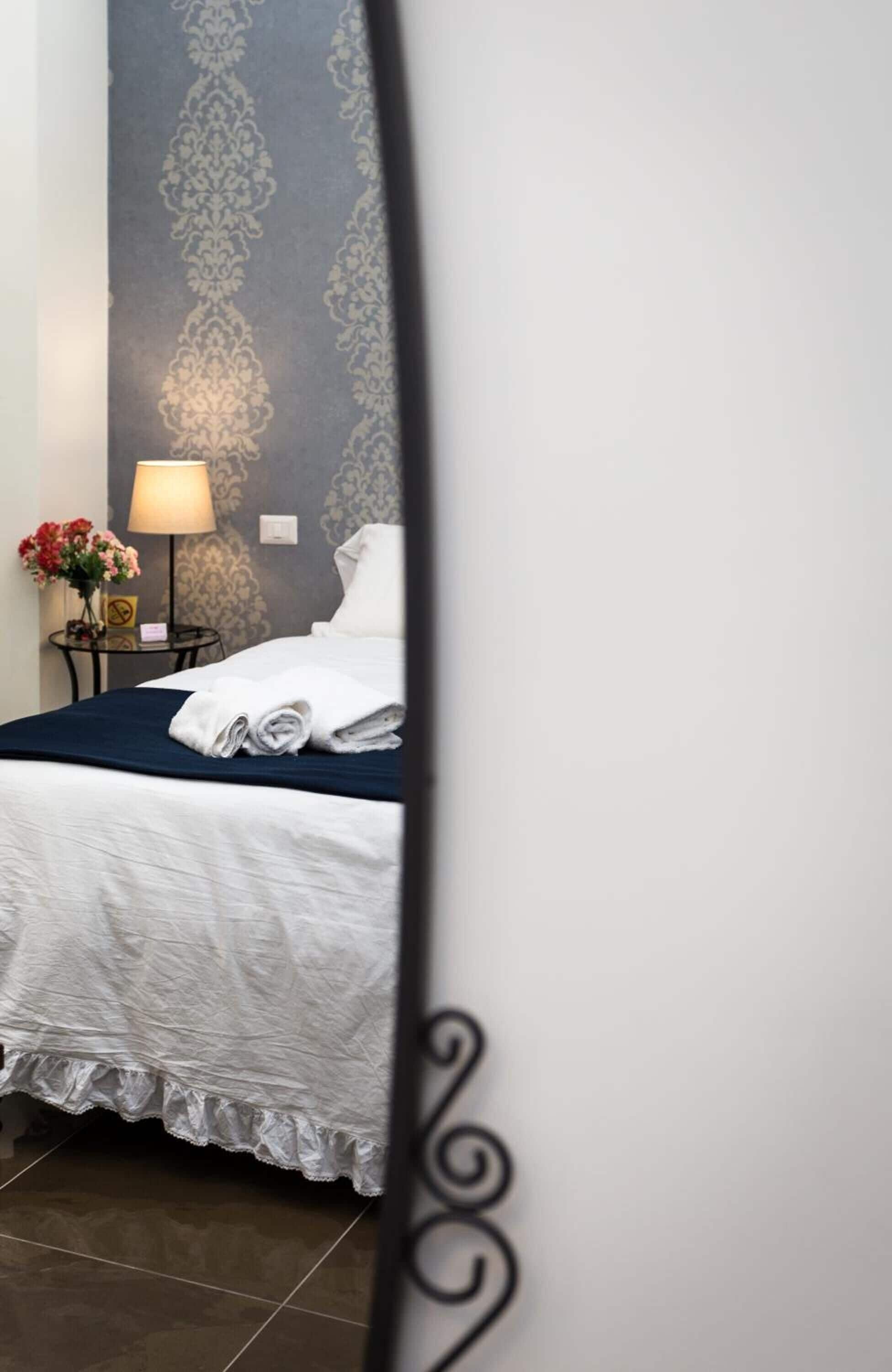 Un lit bien fait, avec des draps blancs et une couvre-lit bleue, une table de chevet avec une lampe et des fleurs, et un papier peint à motifs sur le mur.