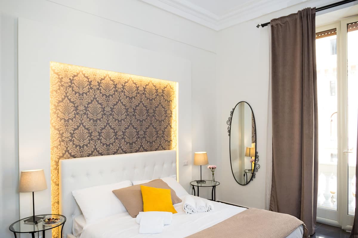 Ett modernt hotellrum med en stor säng, en sänggavel med ett mönstrat motiv, en rund spegel och ett litet bord med en lampa.