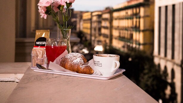 Un balcón con una mesa dispuesta para una comida, que incluye un croissant, una taza con la etiqueta "Caff. Beccucci" y un vaso de bebida roja.