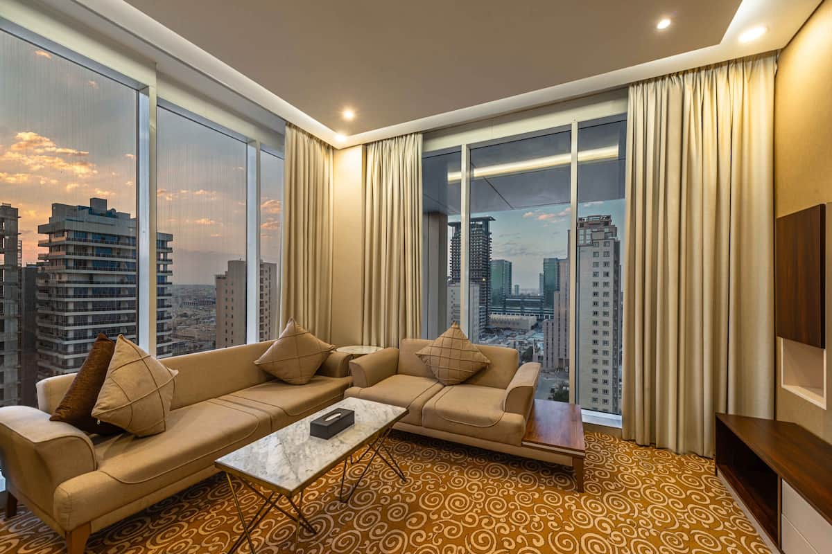 A modern hotel room with a large window, a sofa set, a small coffee table, and a TV on the wall.