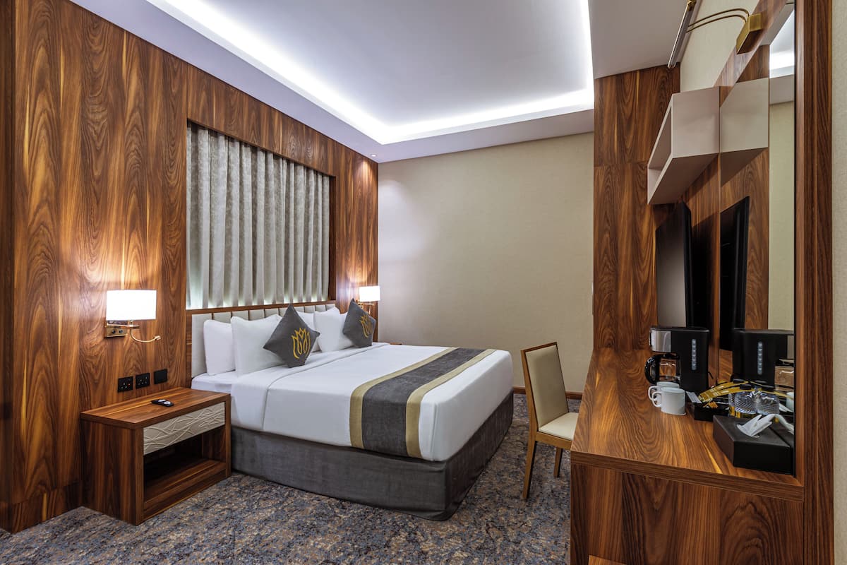 A hotel room with a wooden panel wall, a large bed with white bedding, a bedside table with a lamp, a chair, and a desk with a coffee maker and other amenities.