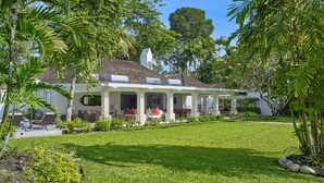 Een tweelaags huis met een overdekte veranda, omgeven door een verzorgd gazon en tropische planten.