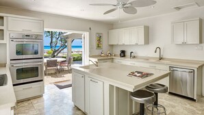 Een moderne keuken met witte kasten, een groot eiland met krukken, een plafondventilator en uitzicht op zee.