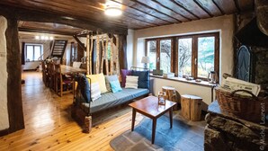 A cozy living room with a wooden ceiling, a sofa with colorful cushions, a dining area with wooden chairs, and a large window with a view of trees.