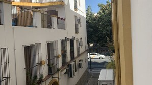 A narrow street with a white building featuring balconies with metal railings and potted plants. - Hostal Alcobia (Seville)