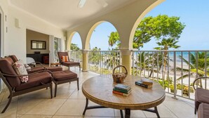A balcony with a view of the beach, furnished with a round table, chairs, and a ceiling fan. - Modern Beachfront Condo with Pool - Sapphire 209 (Saint Lawrence Gap)