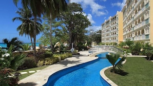 A resort pool with a bridge, surrounded by palm trees and a building with multiple balconies. - Modern Beachfront Condo with Pool - Sapphire 209 (Saint Lawrence Gap)