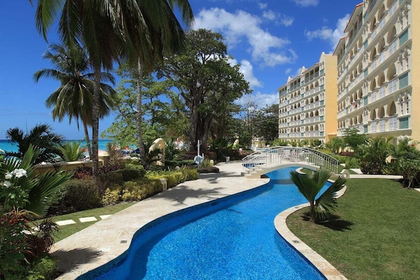 A resort pool with a bridge, surrounded by palm trees and a building with multiple balconies.