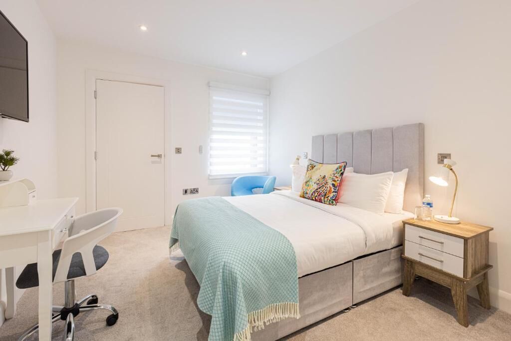 A bedroom with a bed, bedside table, lamp, chair, desk, and a window with blinds.