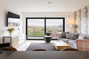 A modern living room with a large window, a flat-screen TV, a sofa, a coffee table, and a dining area. - Curran Gate Luxury Apartments, Portrush (Portrush)