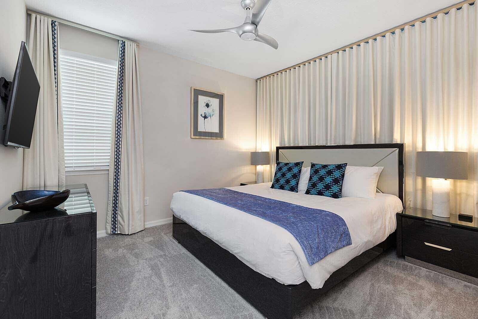 A modern hotel room with a large bed, a flat-screen TV, a ceiling fan, and a view of the city.