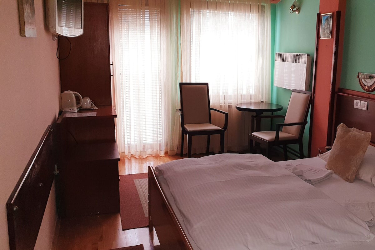 Habitación de hotel con una cama, un escritorio con teléfono, una silla, una mesita y una ventana con cortinas.
