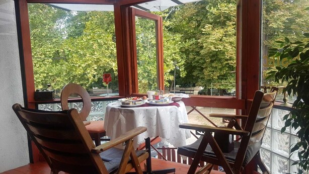 A covered patio with a table set for two, surrounded by wooden chairs, overlooking a garden with trees. - Hotel Kapistec (Skopje)