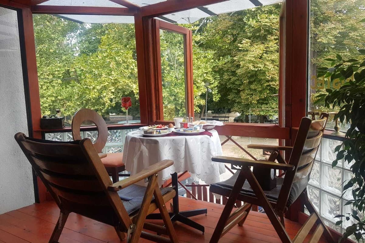 Un patio techado con una mesa para dos, rodeado de sillas de madera, con vista a un jardín con árboles.