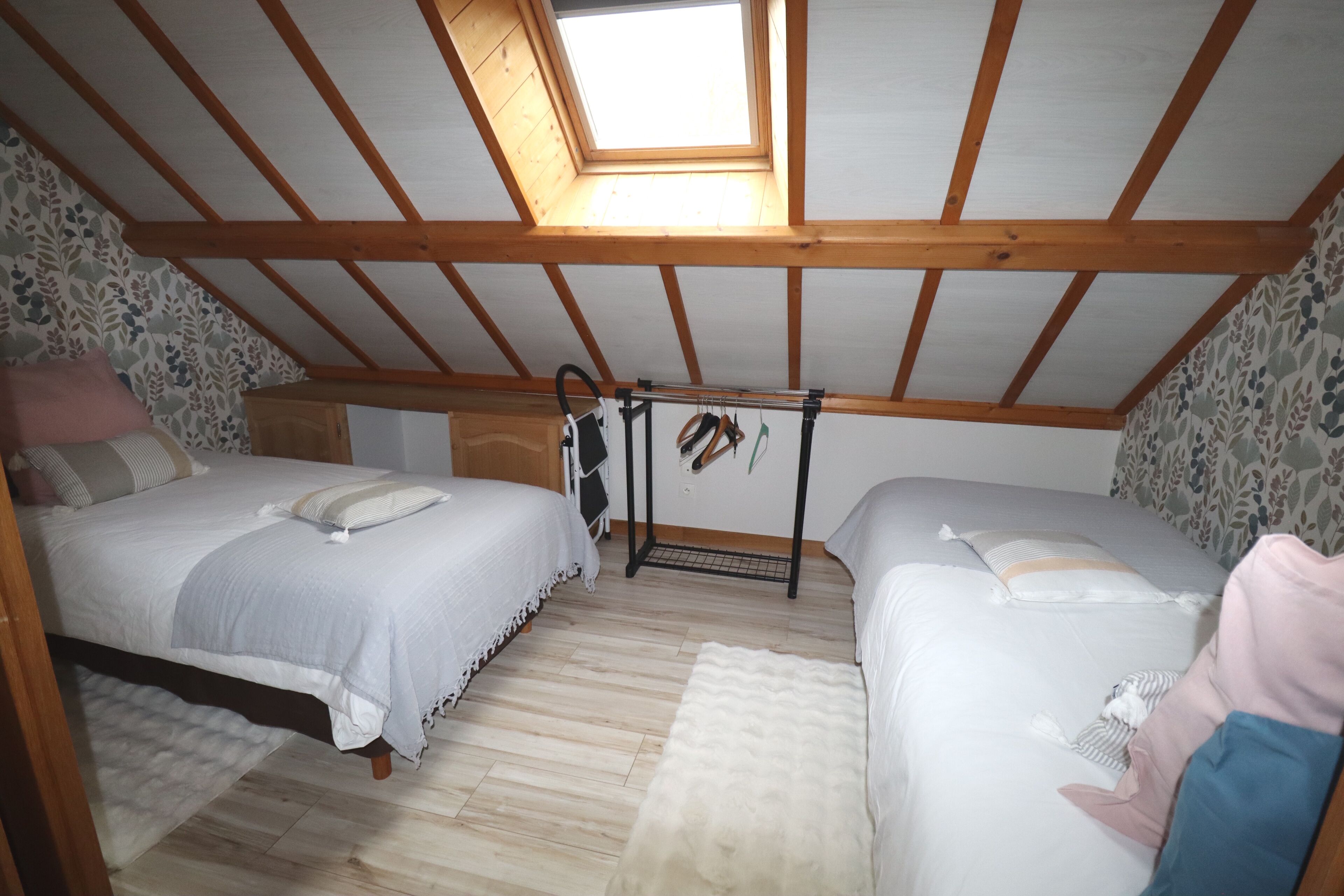 Une chambre avec deux lits, une lucarne, des poutres en bois, un papier peint à motifs et une petite armoire.
