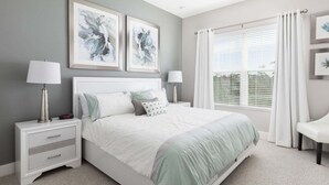 A bedroom with a bed, two bedside tables, a nightstand, a window with curtains, and two framed artworks on the wall.