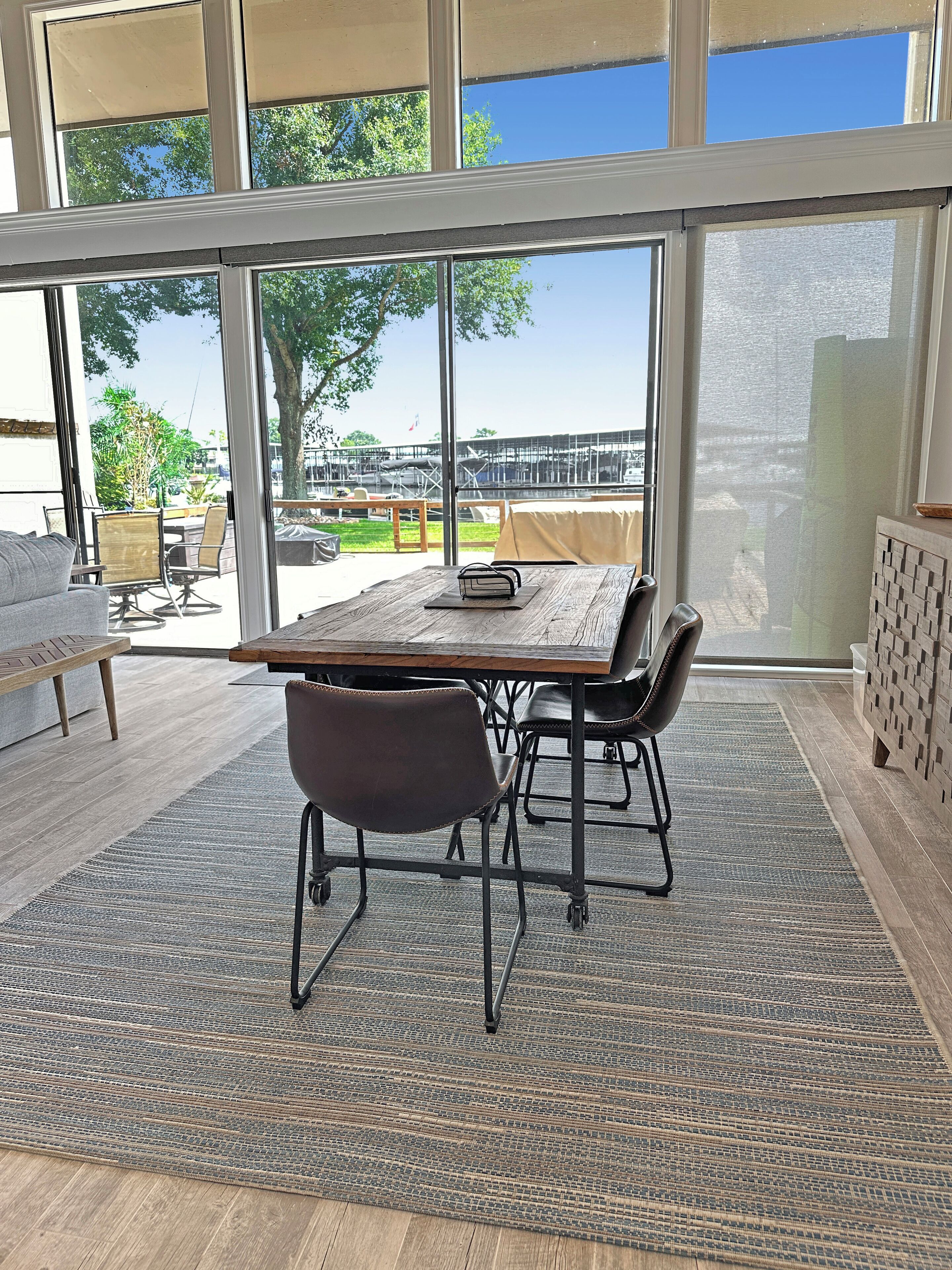 Un comedor moderno con una mesa grande y sillas, ventanas amplias con vista a un puerto deportivo y vegetación.