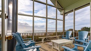 Een overdekt terras met blauwe Adirondack-stoelen, een witte tafel en uitzicht op het strand.