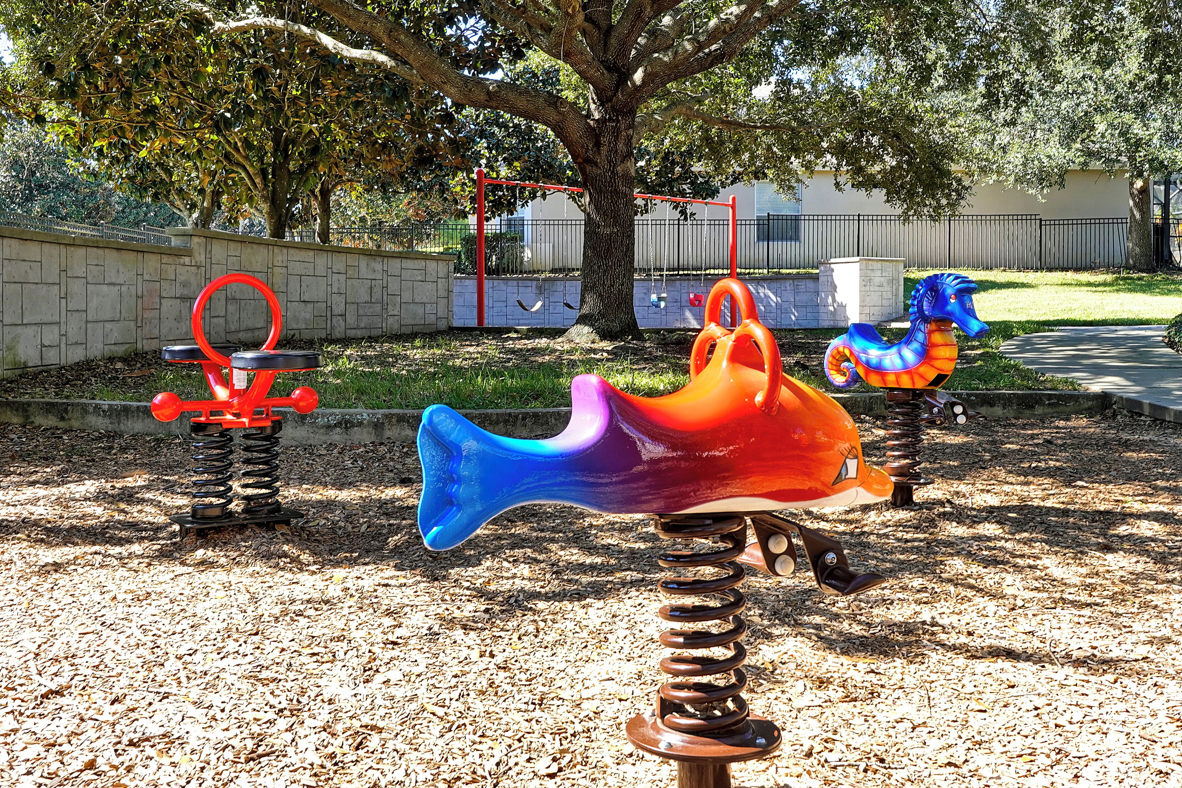 A playground with colorful play equipment, including swings and a slide, set against a backdrop of trees and a fence.