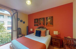 Habitación de hotel con cama, mesita de noche, una silla y un balcón con vistas a edificios y zonas verdes.