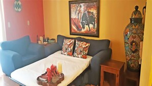 Una habitación con sofá cama, una pequeña mesa de madera, un jarrón decorativo y una obra de arte enmarcada en la pared.
