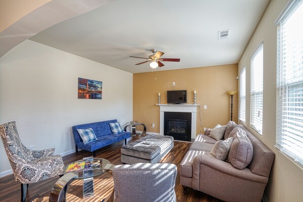 A living room with a blue sofa, a patterned armchair, a grey ottoman, and a beige sofa. There is a fireplace with a mantel and a ceiling fan. - Lovely Long Term/STR Corporate, Family Near I20,Airport,Downtown,Airport (Austell)