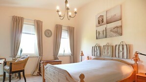 Ein Schlafzimmer mit einem hölzernen Bett, zwei mit Vorhängen versehenen Fenstern, einem Nachttisch, einem Sessel und Wandbildern.