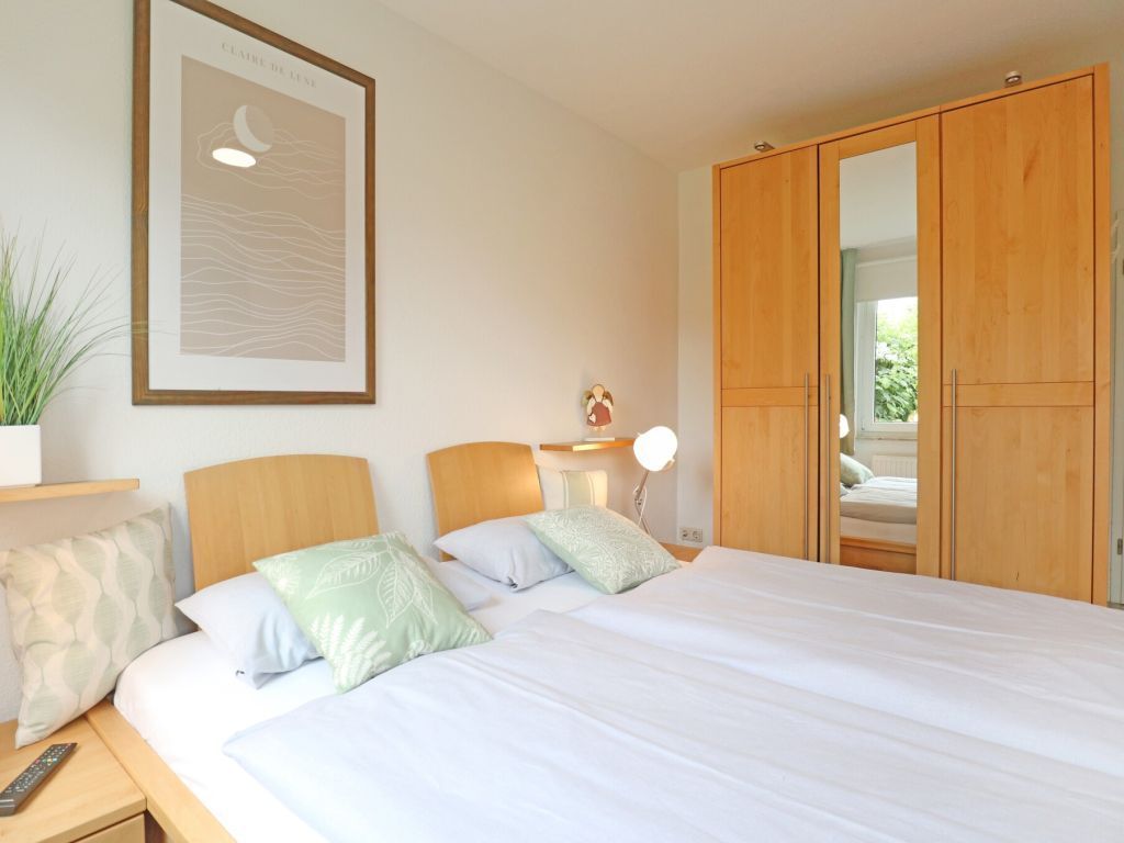 Un dormitorio con una cama grande, armario de madera, mesita de noche con lámpara, una obra de arte enmarcada y una planta en maceta.