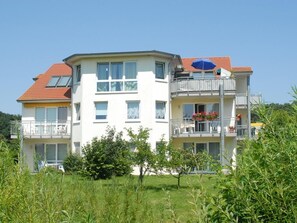 Un immeuble résidentiel à plusieurs étages, doté de balcons, d’un toit recouvert de tuiles rouges et d’un jardin en premier plan.