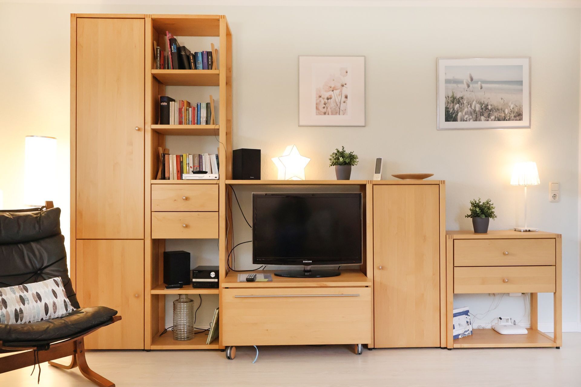 Un salon moderne avec un meuble de rangement en bois pour l’équipement audiovisuel, un canapé en cuir noir, un téléviseur à écran plat et deux lampes fixées au mur.