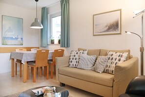 Un salon avec un canapé beige, des coussins à motifs, une table à manger avec des chaises, un luminaire suspendu et un tableau encadré au mur.
