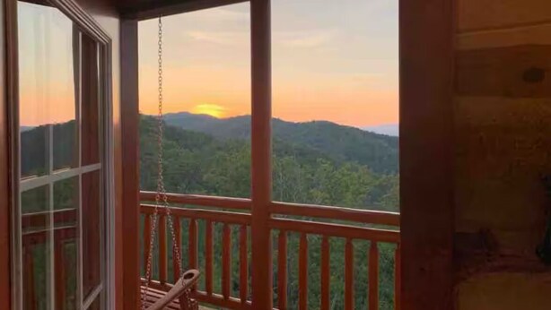 Um deck de madeira com uma cadeira de balanço, uma porta de vidro e vista para um pôr do sol sobre uma cordilheira arborizada.