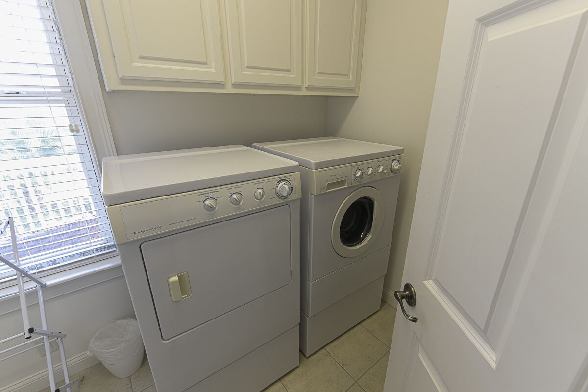 A laundry room with two washing machines and a dryer, white cabinets above, and a window with blinds.