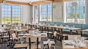 Un restaurante con mesas y sillas de madera, grandes ventanales que ofrecen vistas al exterior y una valla blanca visible a través de las ventanas.