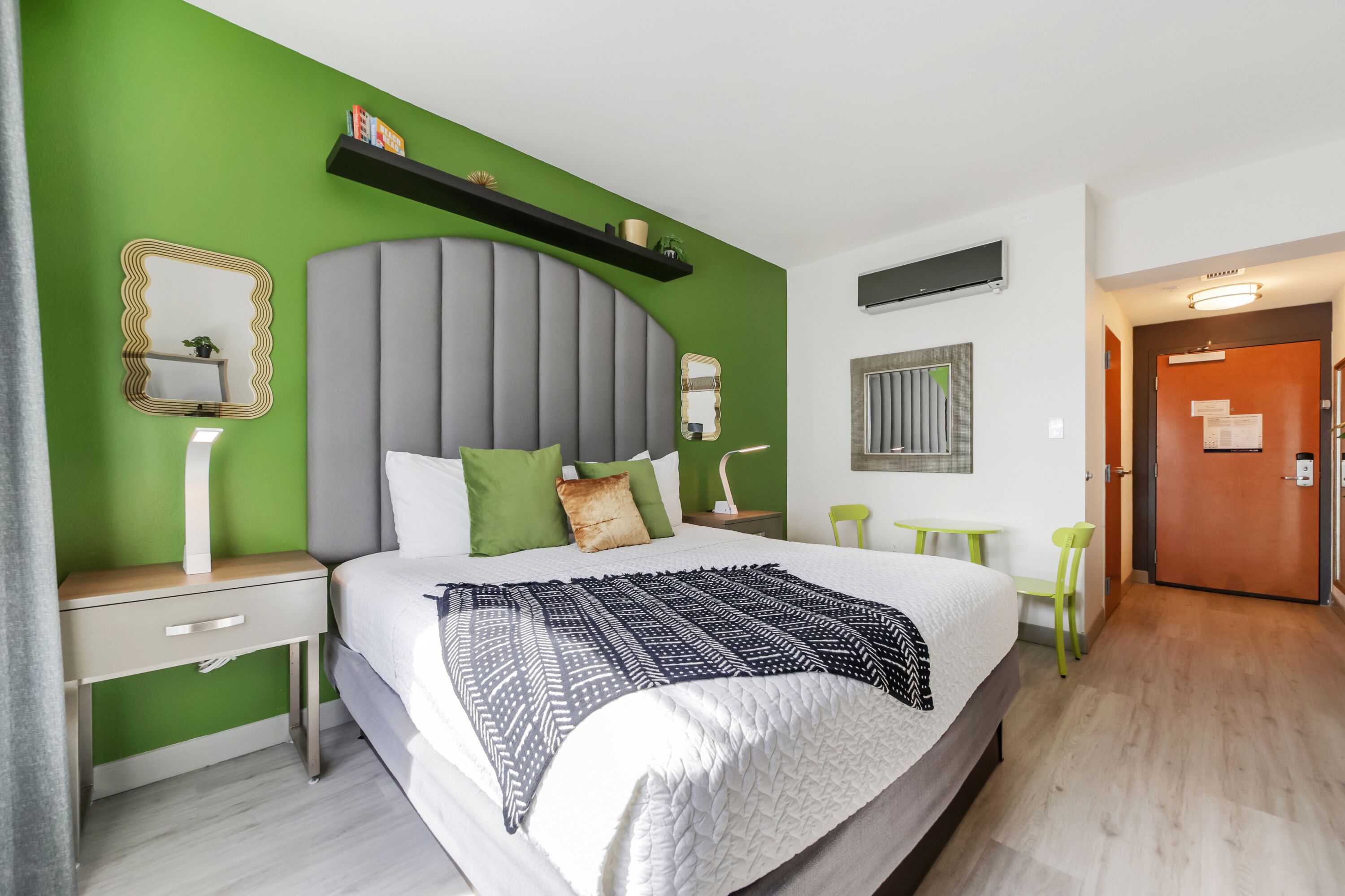 A hotel room with a large bed, green walls, a grey headboard, a bedside table with a lamp, and a small table with chairs.