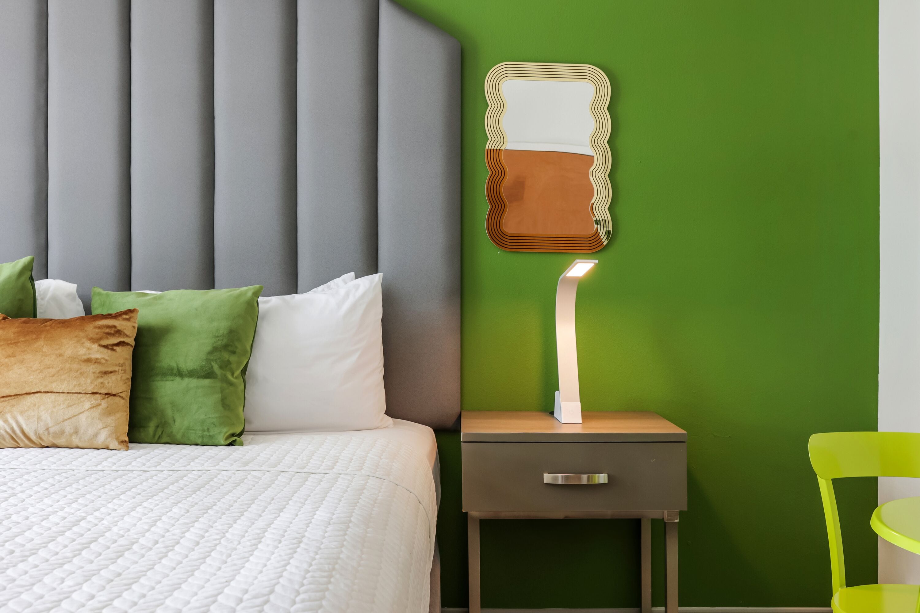 A neatly made bed with white and green pillows, a grey headboard, a bedside table with a drawer, and a green wall.