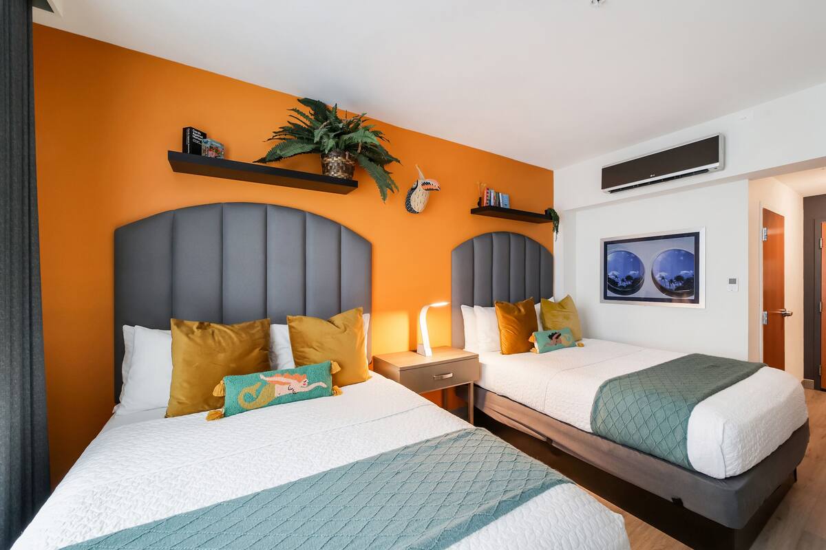 A hotel room with two beds, a large potted plant, and a wall-mounted air conditioner.