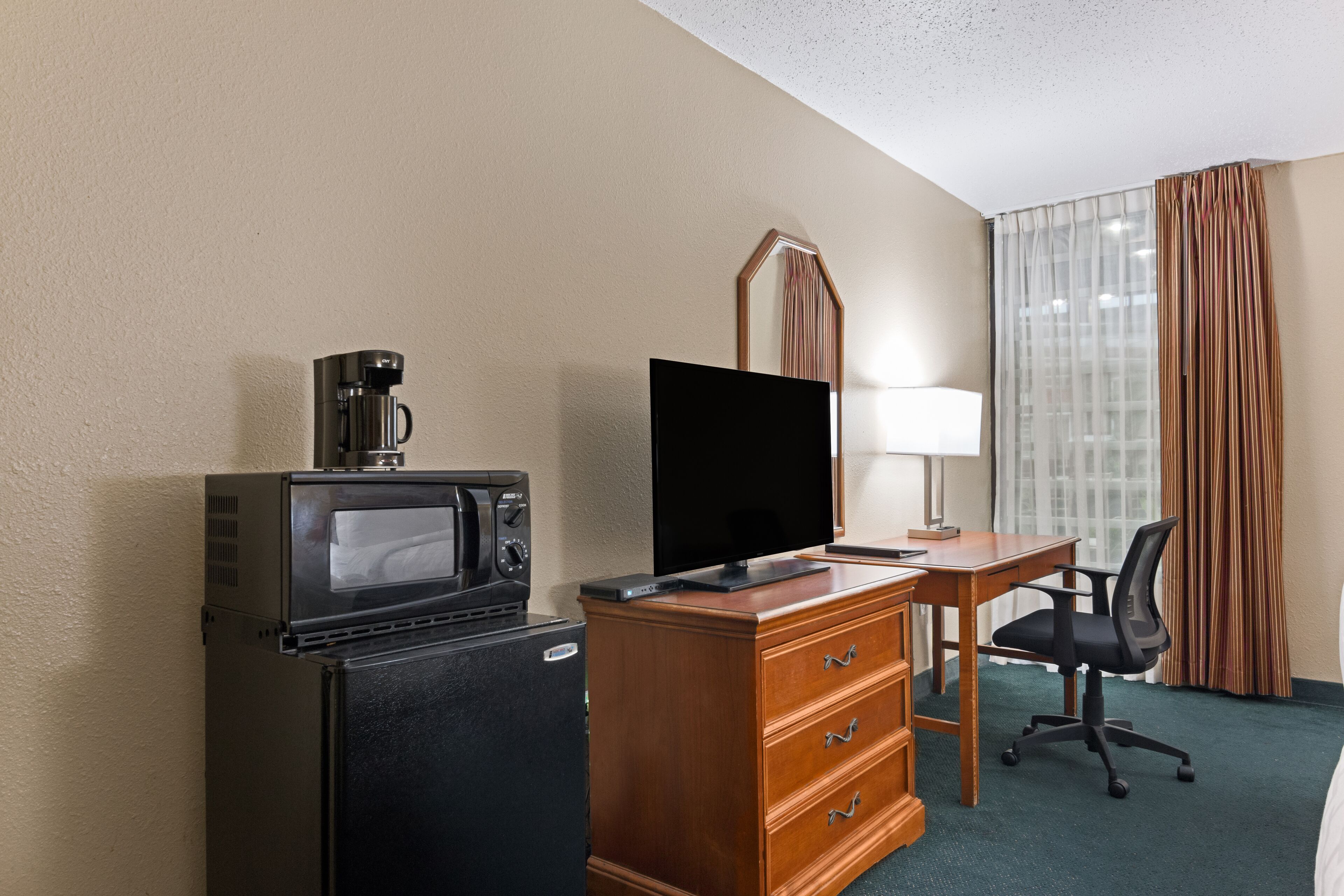 A hotel room with a kitchenette, a flat-screen TV, a wooden dresser, a desk, a chair, and a window with curtains.
