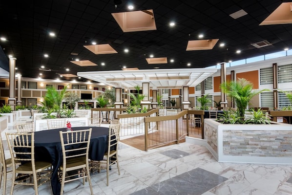A spacious indoor dining area with multiple tables and chairs, surrounded by potted plants and a modern ceiling design.