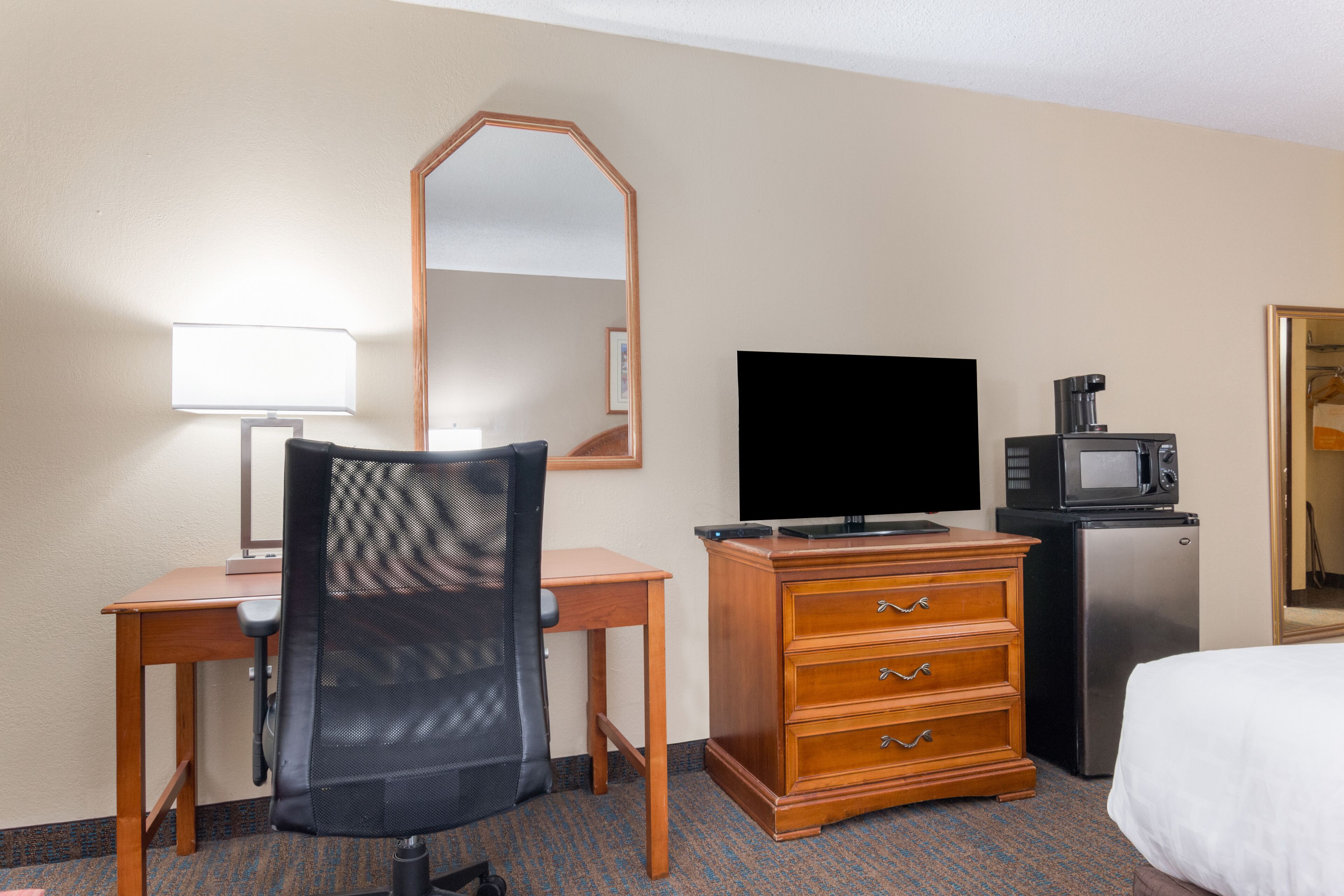 Habitación de hotel con escritorio de madera, una silla de oficina negra, televisor de pantalla plana, microondas, refrigerador, cómoda de madera y cama con ropa blanca.