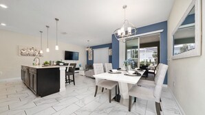 A modern kitchen with a central island, dining area with a white table and chairs, and a large window providing a view of the outdoors. - Elegant 4 Bedroom Getaway w/Private Pool 114 (ChampionsGate)