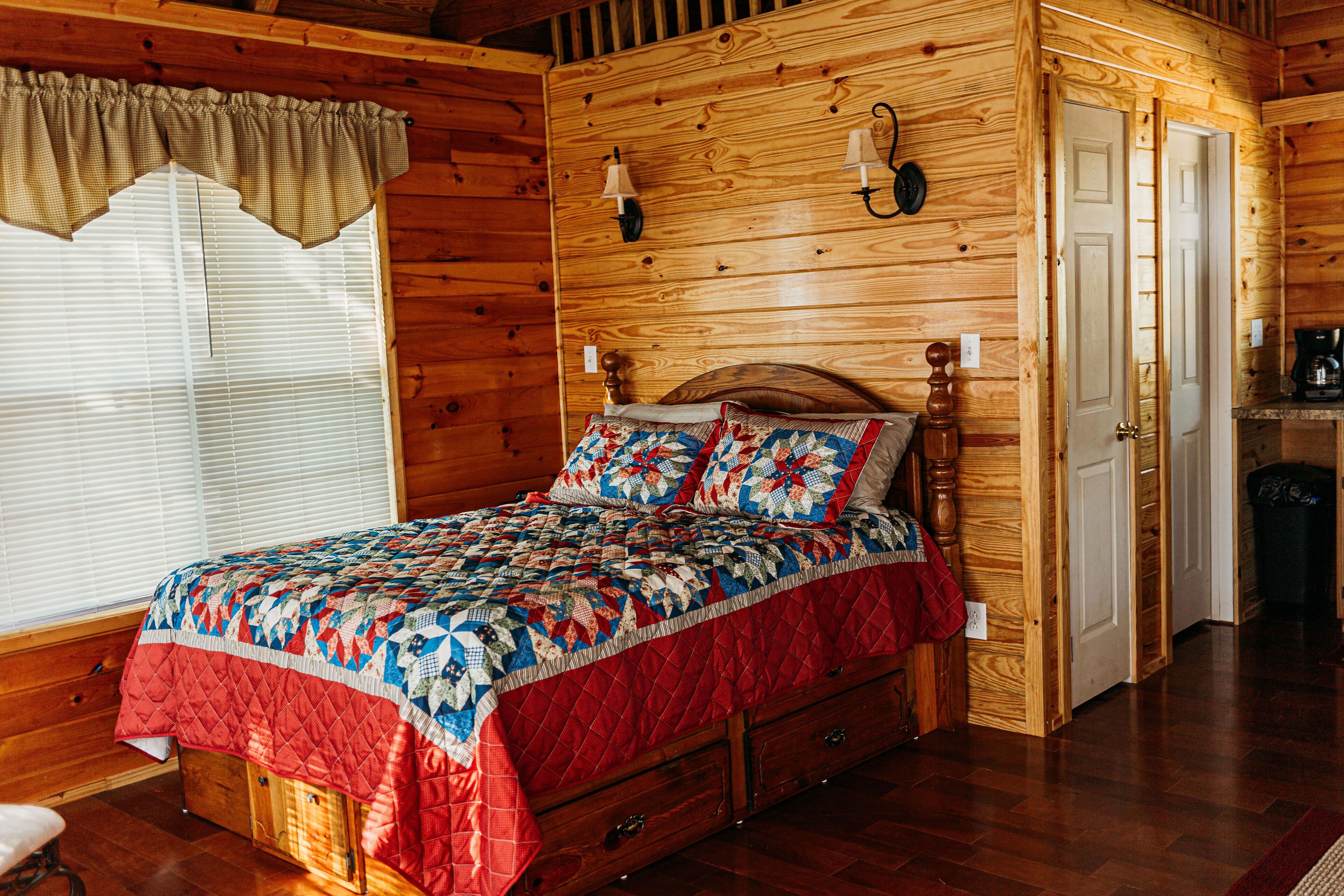 Un dormitorio de cabaña de madera con cama acolchada, paredes de madera, una ventana con persianas y una puerta que da a otra habitación.