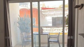 Une chambre avec un lit, une porte-fenêtre coulissante donnant sur un espace extérieur aménagé avec des chaises, une plante en pot et une enseigne indiquant un commerce.